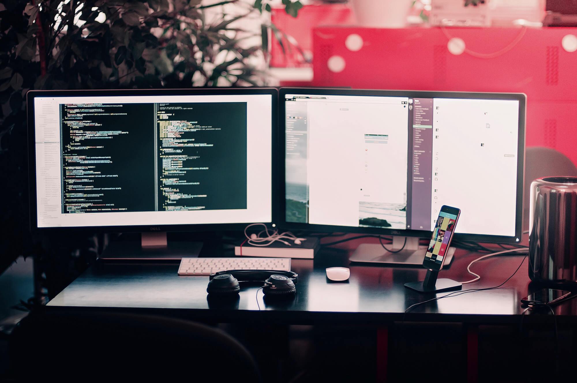 Photograph of a work desk with two monitors displaying a code editor.
