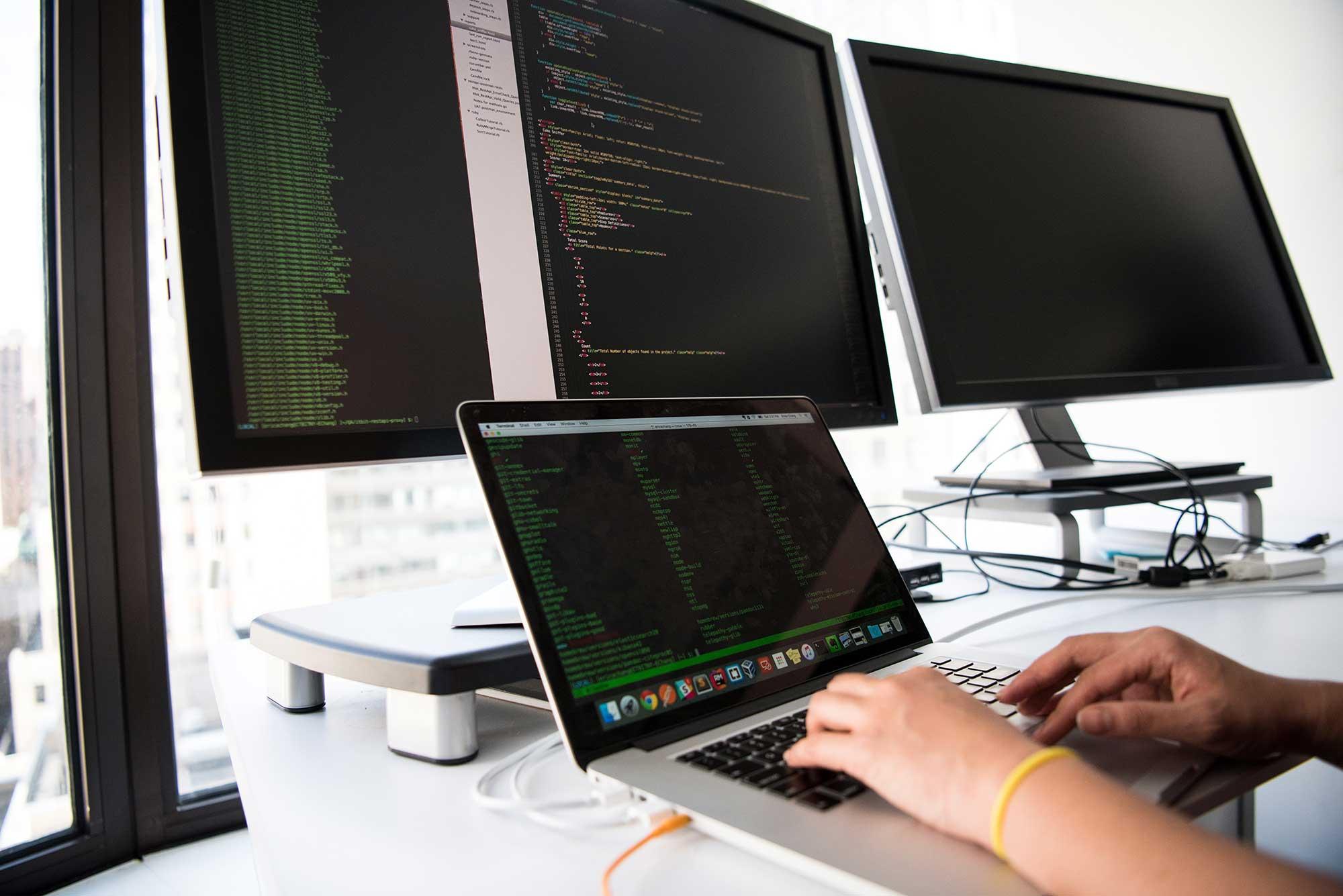 A work desk with multiple screens, each displaying a code editor, as a person types
