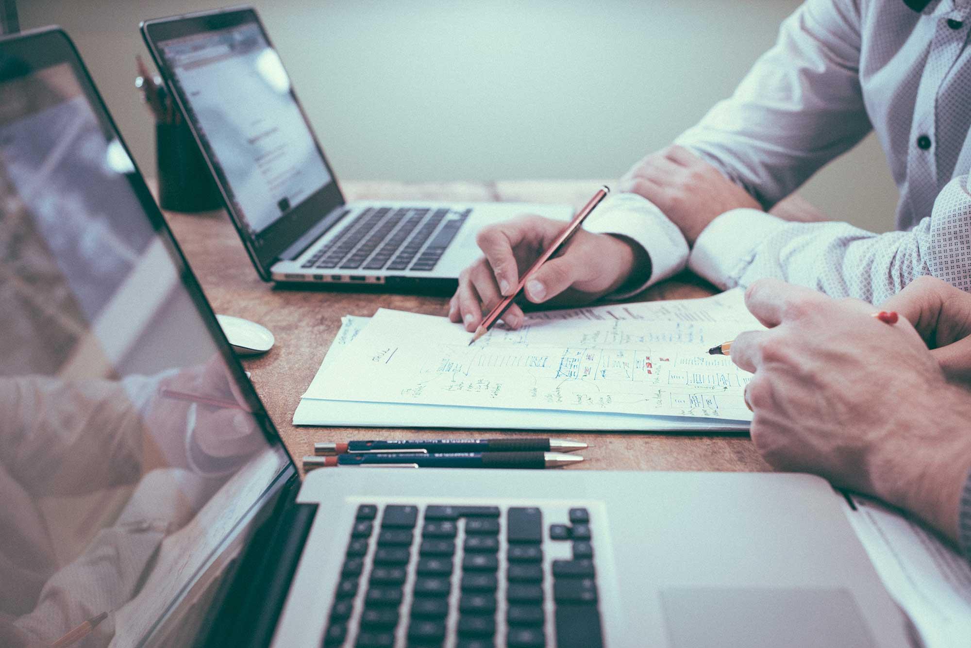 A desk with two laptops and two individuals at them, both collaborating over a piece of paper outlining a dashboard design
