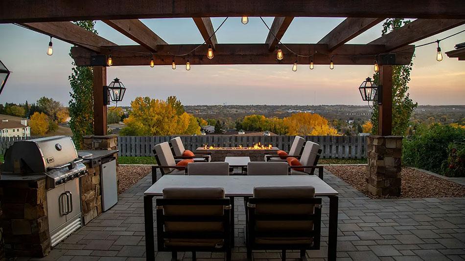 Image of a patio right before sunset. String lights hang from the pergola above a table and a grilling setup. In the background, a firepit is lit invitingly, lending the scene a cozy, welcoming feel.