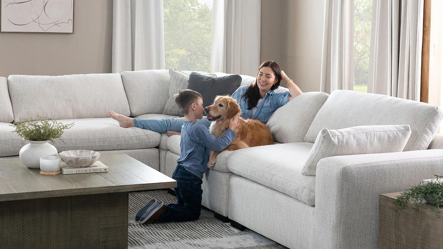 Photograph of a mother and child; the mother lounges on a plush, clean white couch, smiling while her son plays with their golden retriever. The interior decor is minimalistic, modern, and welcoming.