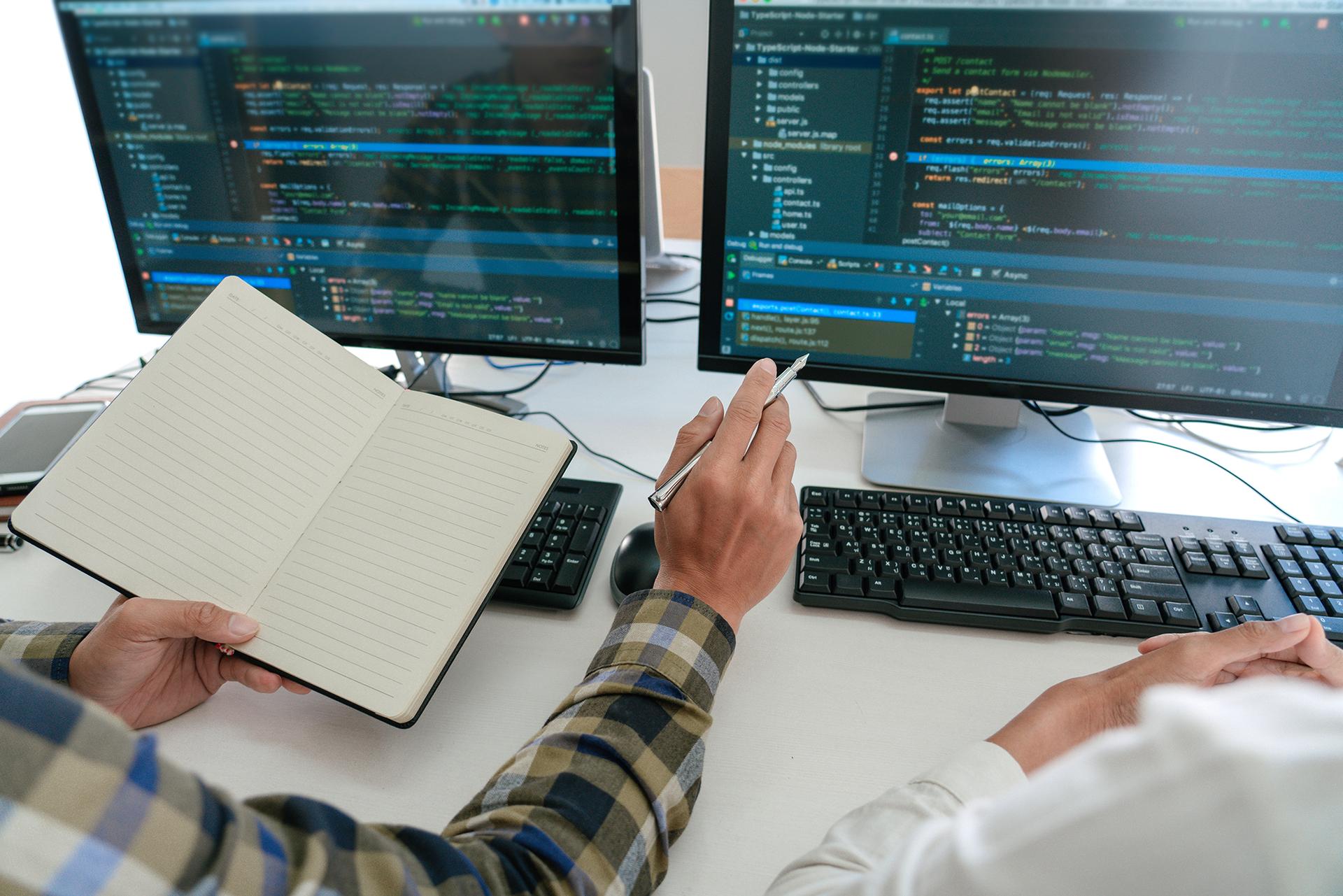 Photograph of two people working on a computer; a code editor is open on both screens.