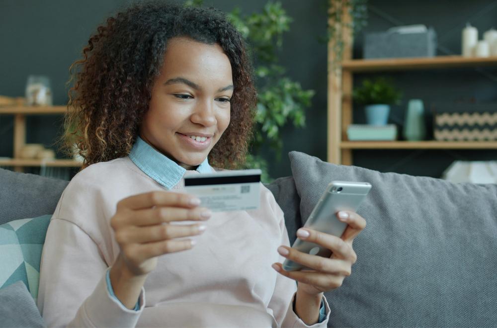 Photograph of a woman sitting on her couch, shopping on her phone with her credit card in the other hand.