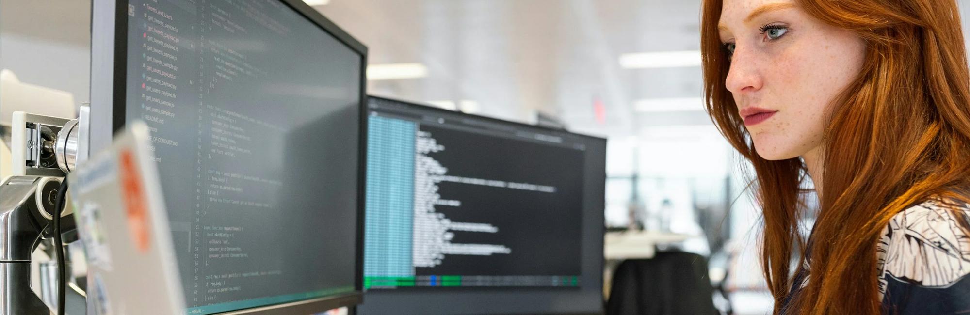 A photograph of a woman in front of a few computer screens, typing and working on a project.